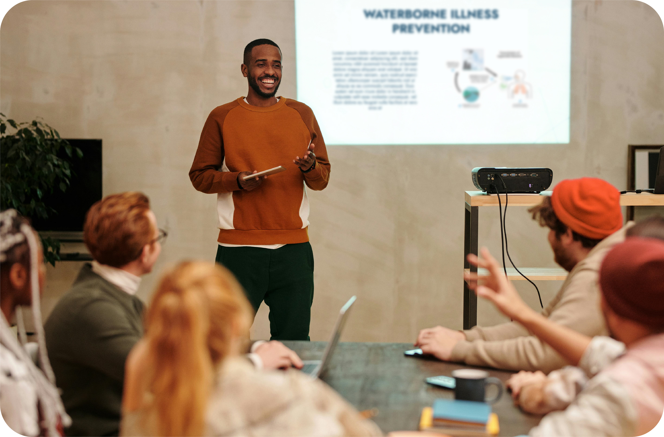 An image of a Black man smiling presenting to a group of people with a blurred projected slide behind him.